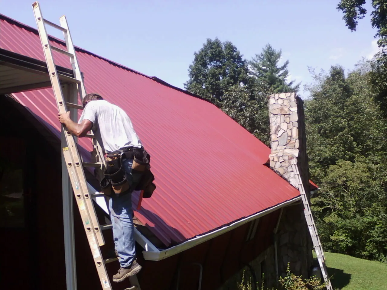 Roofer on a ladder beside a red metal roof and stone chimney — Soffit & Fascia Repair by Roofing Brothers in Acushnet